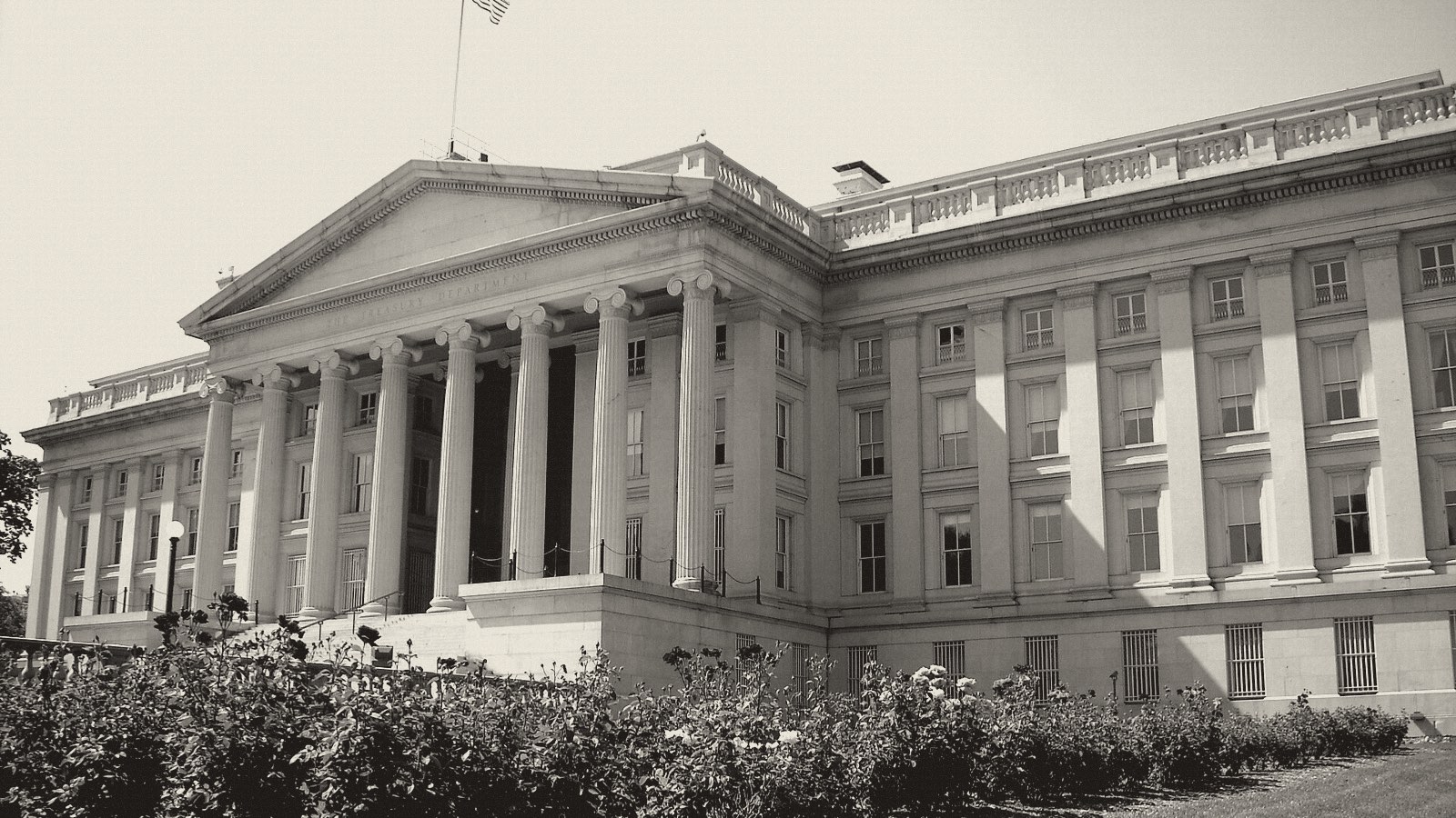 Black-and-white photo of the columned US Treasury Building in Washington, seen at an angle behind a bed of rose bushes.